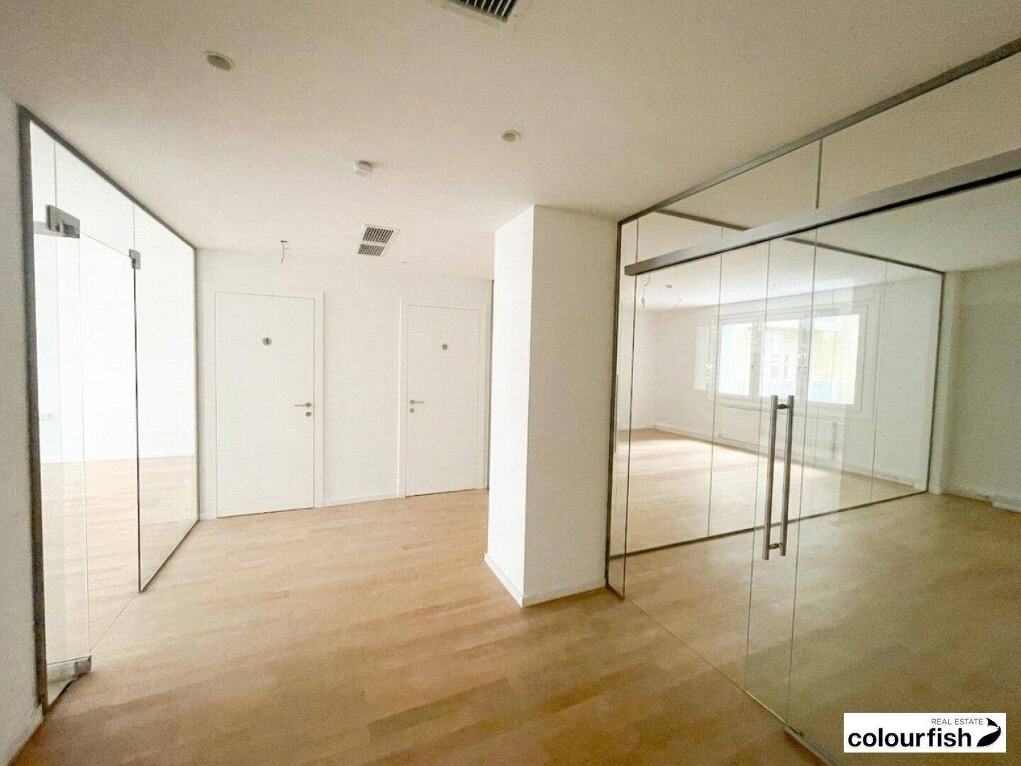 Moderner Bürobereich mit Glaswänden, Holzboden und weißen Türen, der viel Licht hereinlässt.