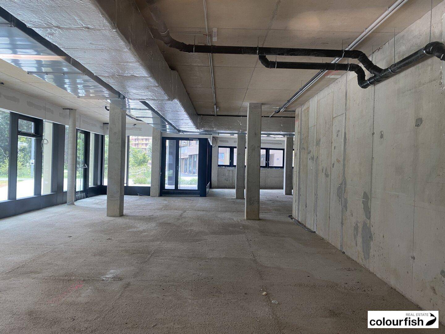 Großer, unfertiger Gewerberaum im Rohbauzustand mit Betonboden, Säulen und sichtbaren Installationen an der Decke.