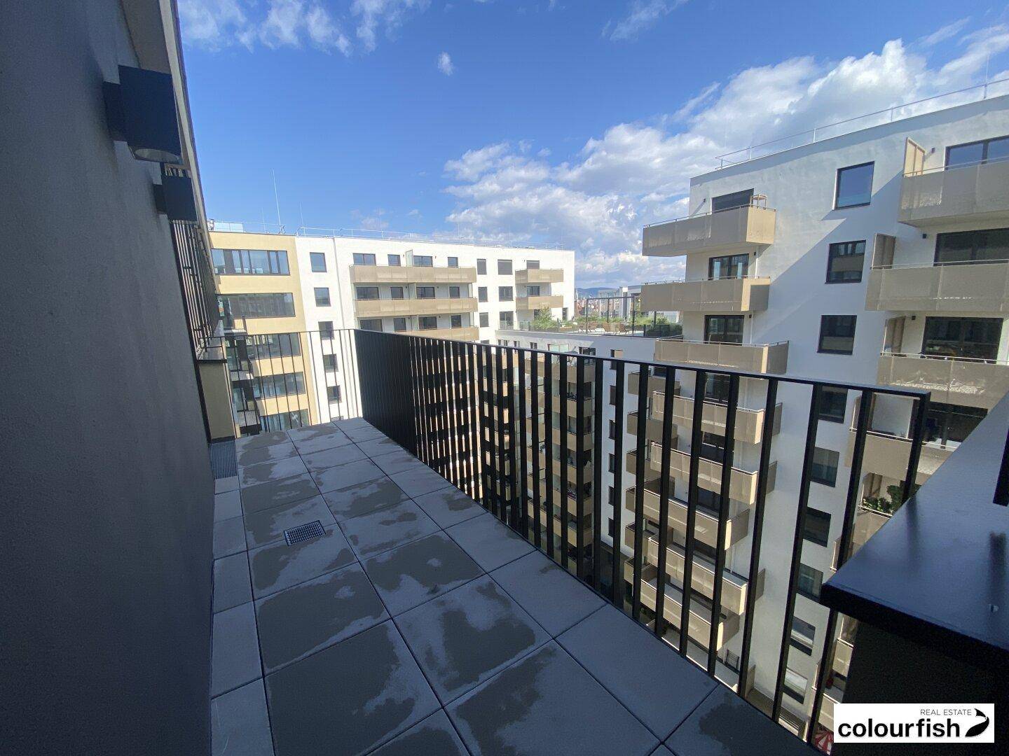 Geräumiger Balkon mit Fliesenboden und Blick auf moderne Wohngebäude unter blauem Himmel.