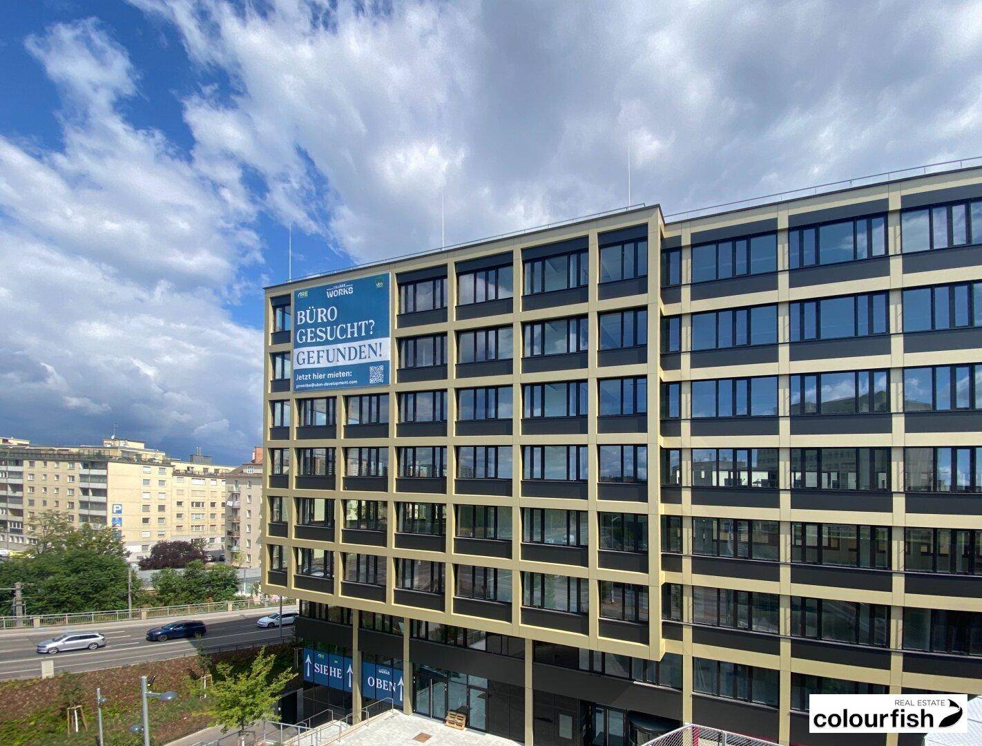 Moderne Gebäudefassade mit großen Fenstern und einer Werbetafel unter blauem Himmel.