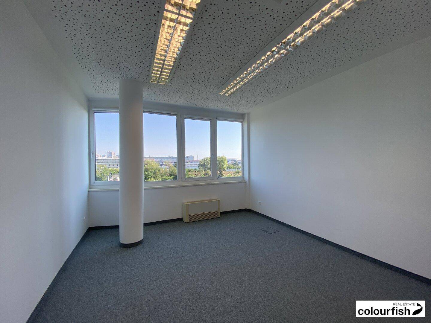 Leerer Büroraum mit Säule, grauem Teppichboden und Fenstern, die einen Blick auf die Stadt und Bäume bieten.