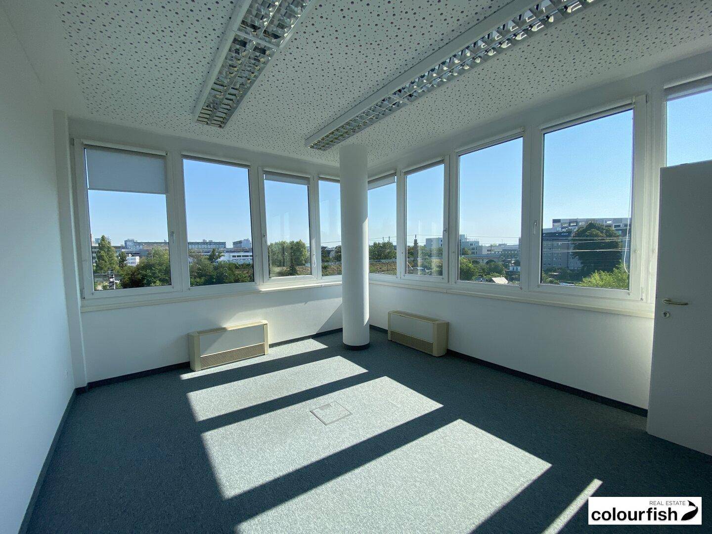 Heller Büroraum in der Ecke mit grauem Teppichboden und vielen Fenstern, die einen weiten Stadtblick ermöglichen.