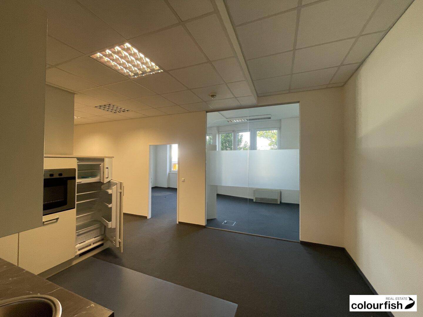 Kleiner Küchenbereich mit Einbaukühlschrank und Ofen, angrenzend an einen Büroraum mit Glastrennwand.