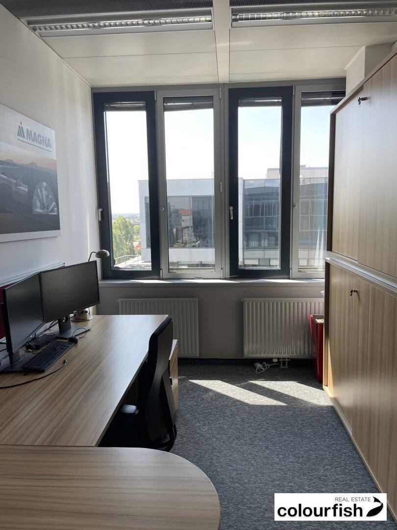 Kleines Büro mit Schreibtisch, Computermonitoren und einem großen Fenster, das viel Tageslicht hereinlässt.