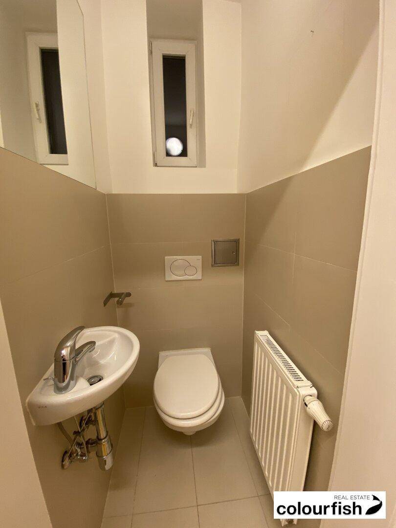 Modernes Gäste-WC mit wandhängender Toilette, kleinem Waschbecken und einem schmalen Fenster für natürliche Belüftung.