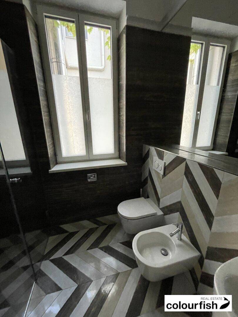 Modernes Badezimmer mit auffälligen Chevron-Fliesen, wandhängendem WC, Bidet und einem Fenster für Tageslicht.