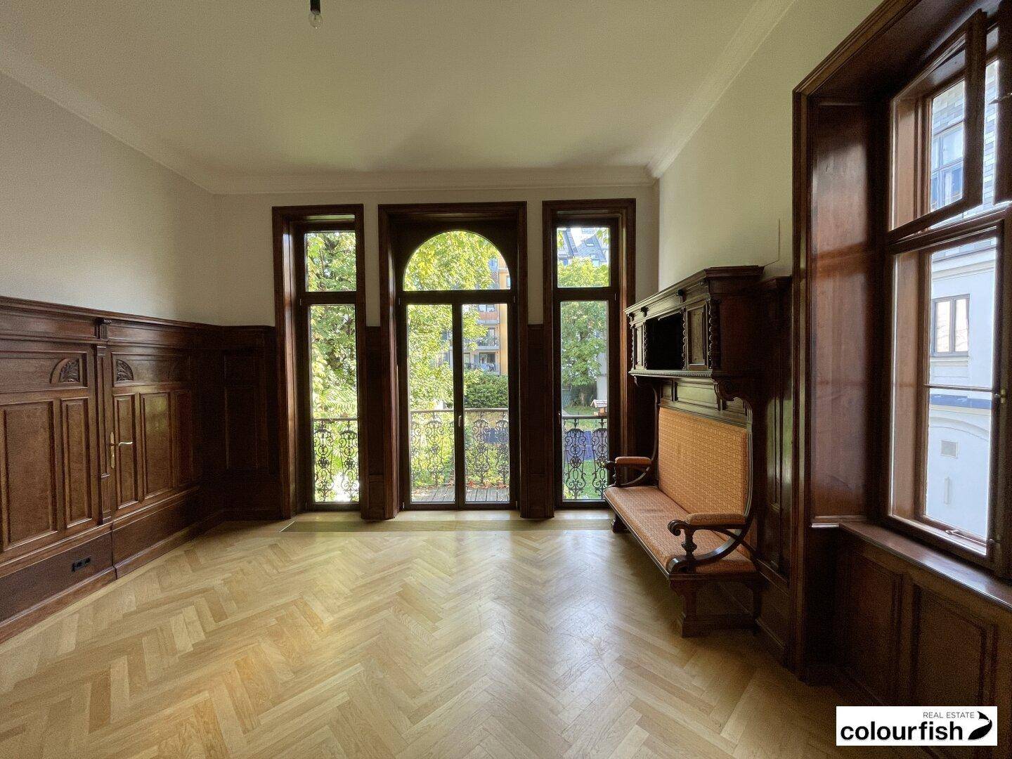 Heller Raum mit Fischgrätparkett, Holzvertäfelung und hohen Fenstertüren, die Zugang zu einem kleinen Balkon bieten.