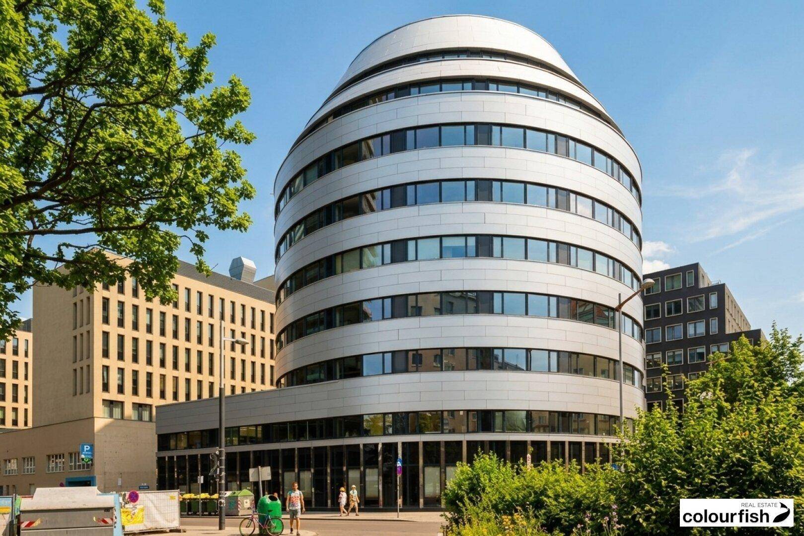 Modernes, rundes Bürogebäude mit markanter Fassade und vielen Fenstern unter blauem Himmel.