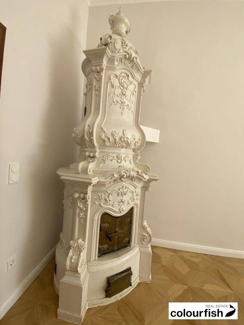 Prunkvoller weißer Kachelofen mit reichen Verzierungen in einem Raum mit elegantem Parkettboden.