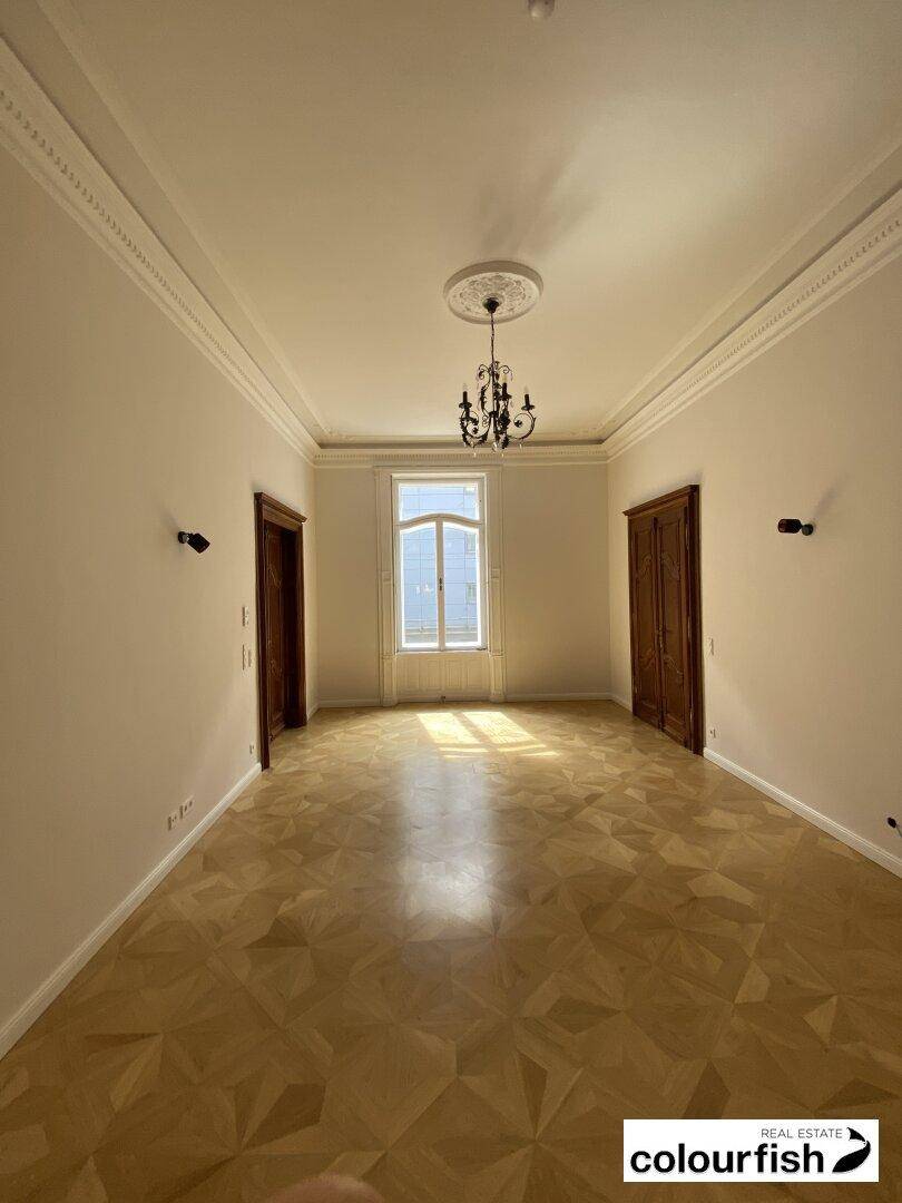 Heller, leerer Raum mit klassischem Parkettboden, einem großen Fenster und einem eleganten Kronleuchter.
