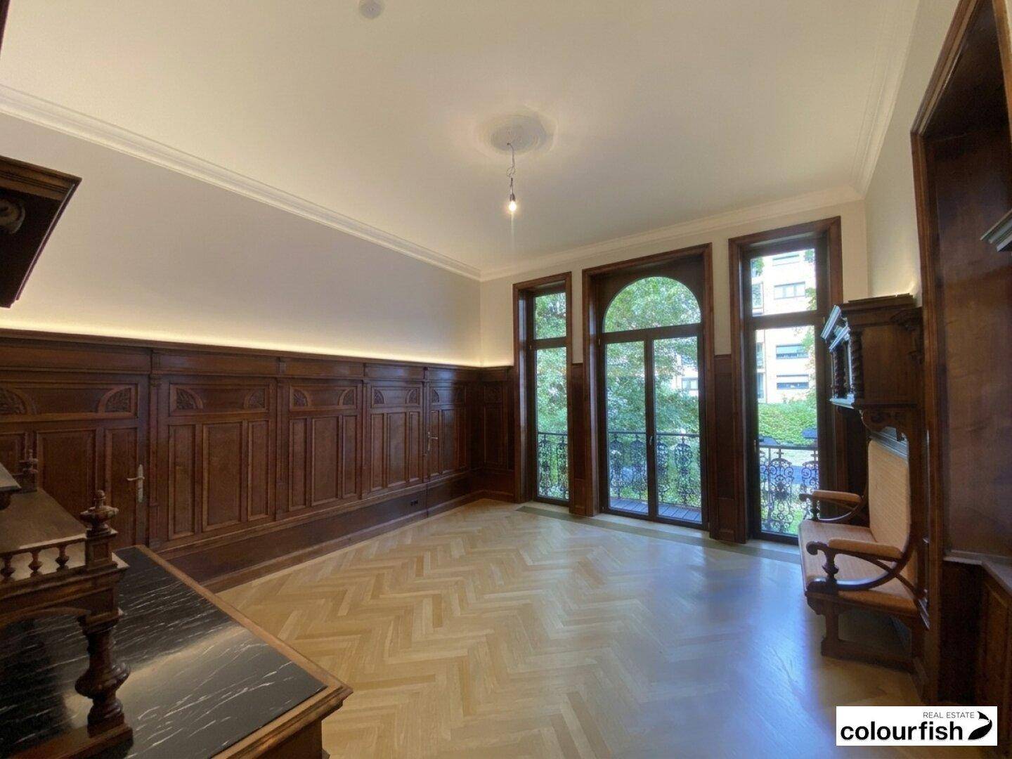 Geräumiges Wohnzimmer mit dunkler Holzvertäfelung, Fischgrätparkett und hohen Fenstern mit Blick ins Grüne.