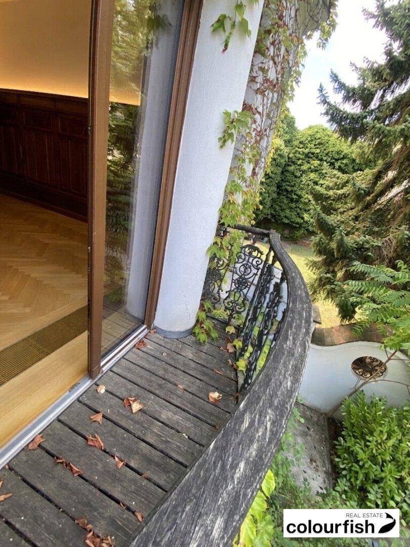 Kleiner, geschwungener Holzbalkon mit schmiedeeisernem Geländer und Blick auf einen üppigen Garten.