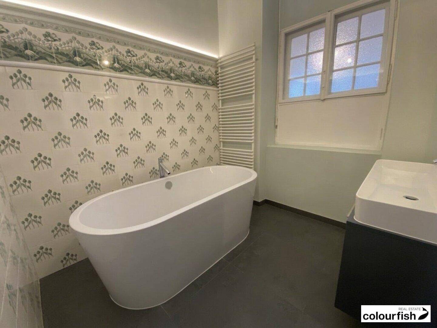 Modernes Badezimmer mit freistehender Badewanne, stilvollem Waschtisch, Fenster und gemusterten Fliesen.