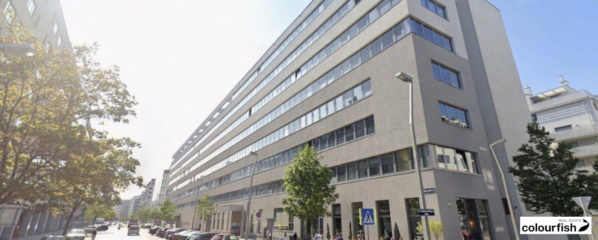 Modernes Bürogebäude mit vielen Fenstern und klaren Linien, umgeben von Bäumen an einer Stadtstraße.