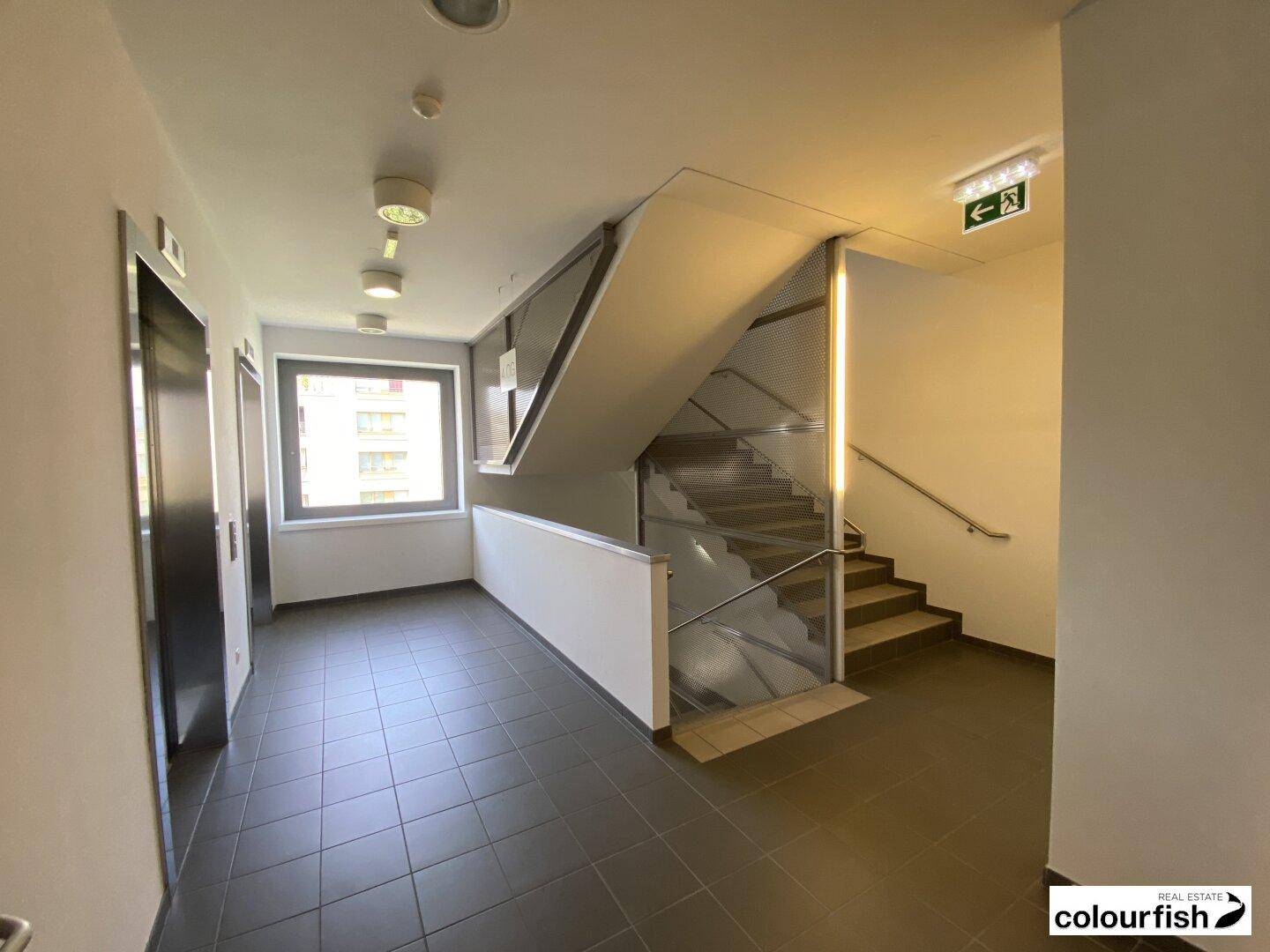 Heller Treppenhausbereich mit Fliesenboden und einem Fenster, das Tageslicht hereinlässt, neben einem Aufzug.