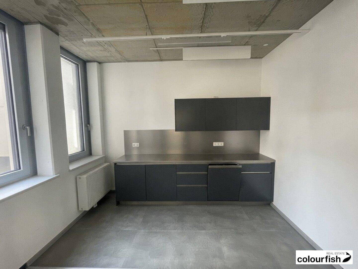 Moderne Küchenzeile mit dunklen Schränken, Edelstahl-Arbeitsplatte und Fliesenboden in einem Büroraum.
