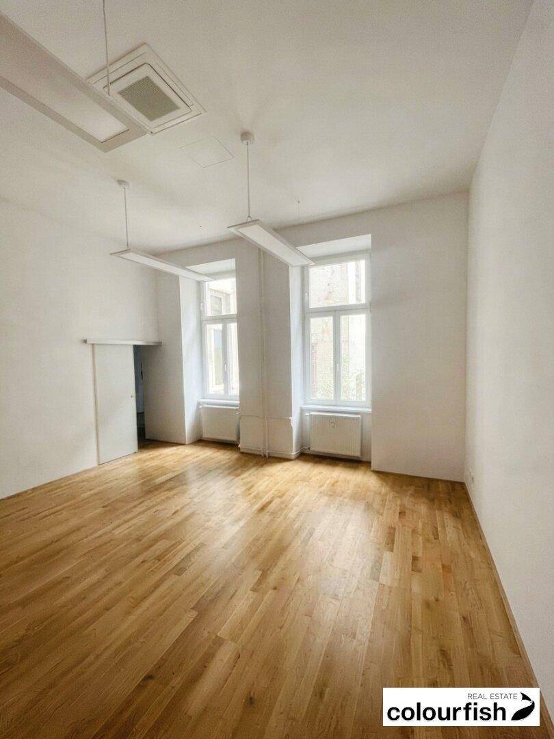 Heller Raum mit Holzboden, hohen Fenstern und moderner Deckenbeleuchtung, ideal als Büro.