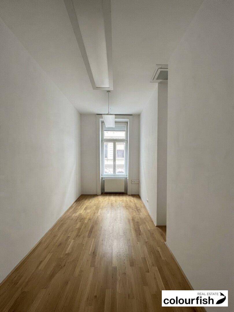 Schmaler, heller Raum mit Holzboden und einem großen Fenster, geeignet als kleines Büro oder Flur.
