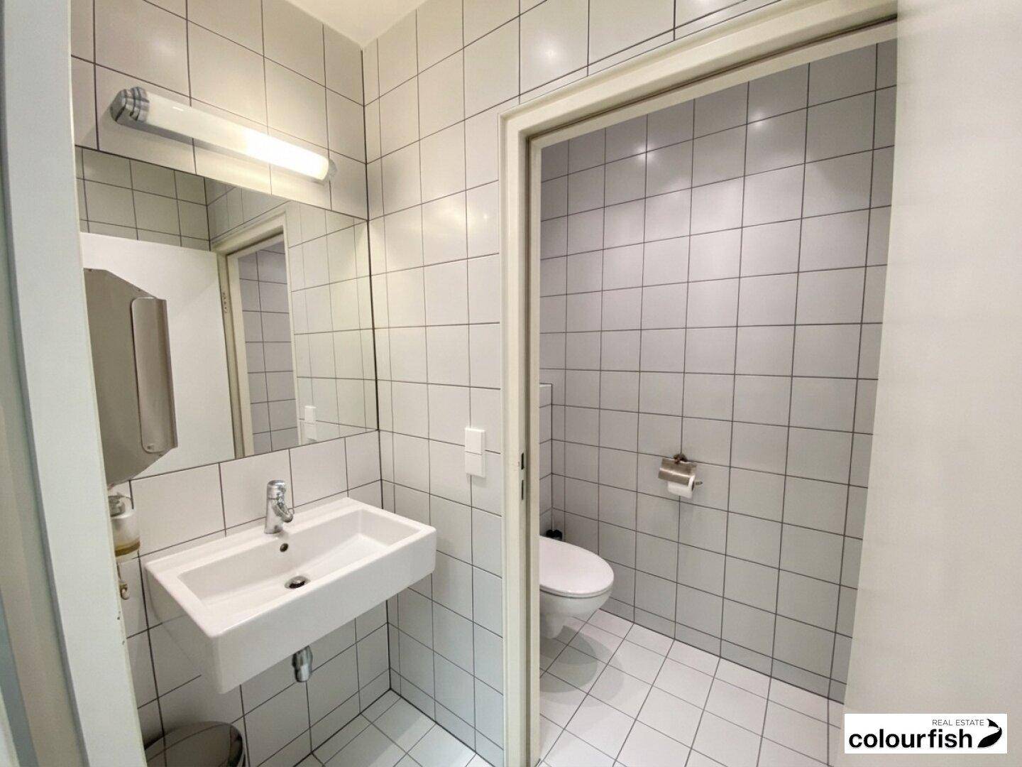 Modernes Badezimmer mit weißen Fliesen, wandmontiertem Waschbecken und separatem WC-Bereich.