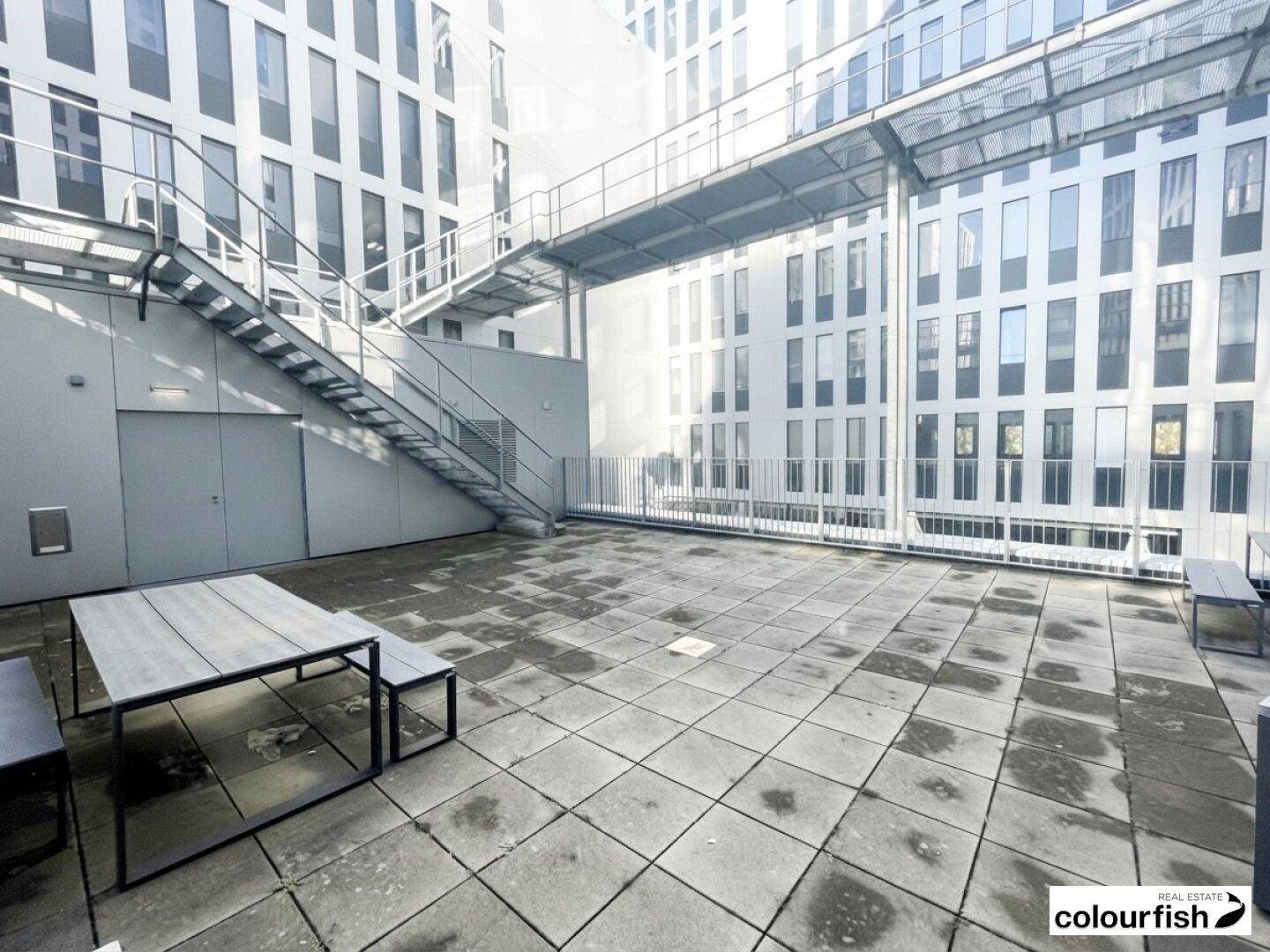 Große Außenterrasse mit Steinplatten, Tischen und Bänken, umgeben von modernen Gebäuden.