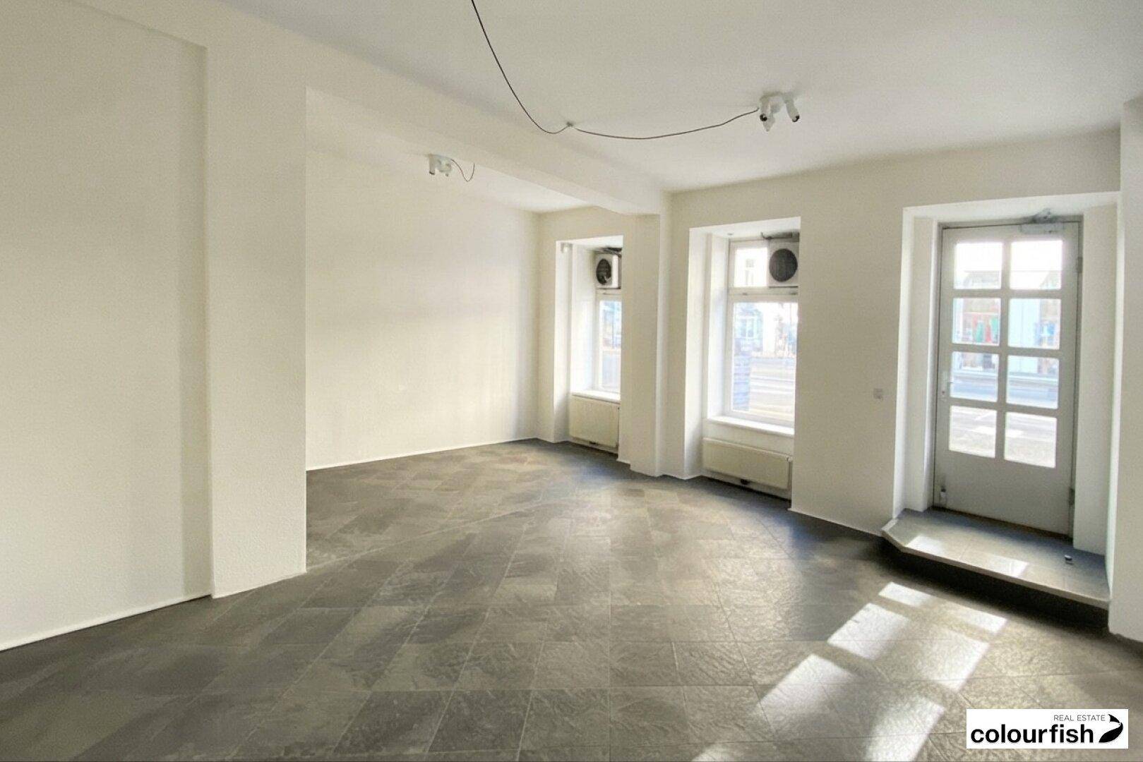 Heller, leerer Geschäftsraum mit großen Fenstern, einer Glastür und dunklem Fliesenboden.