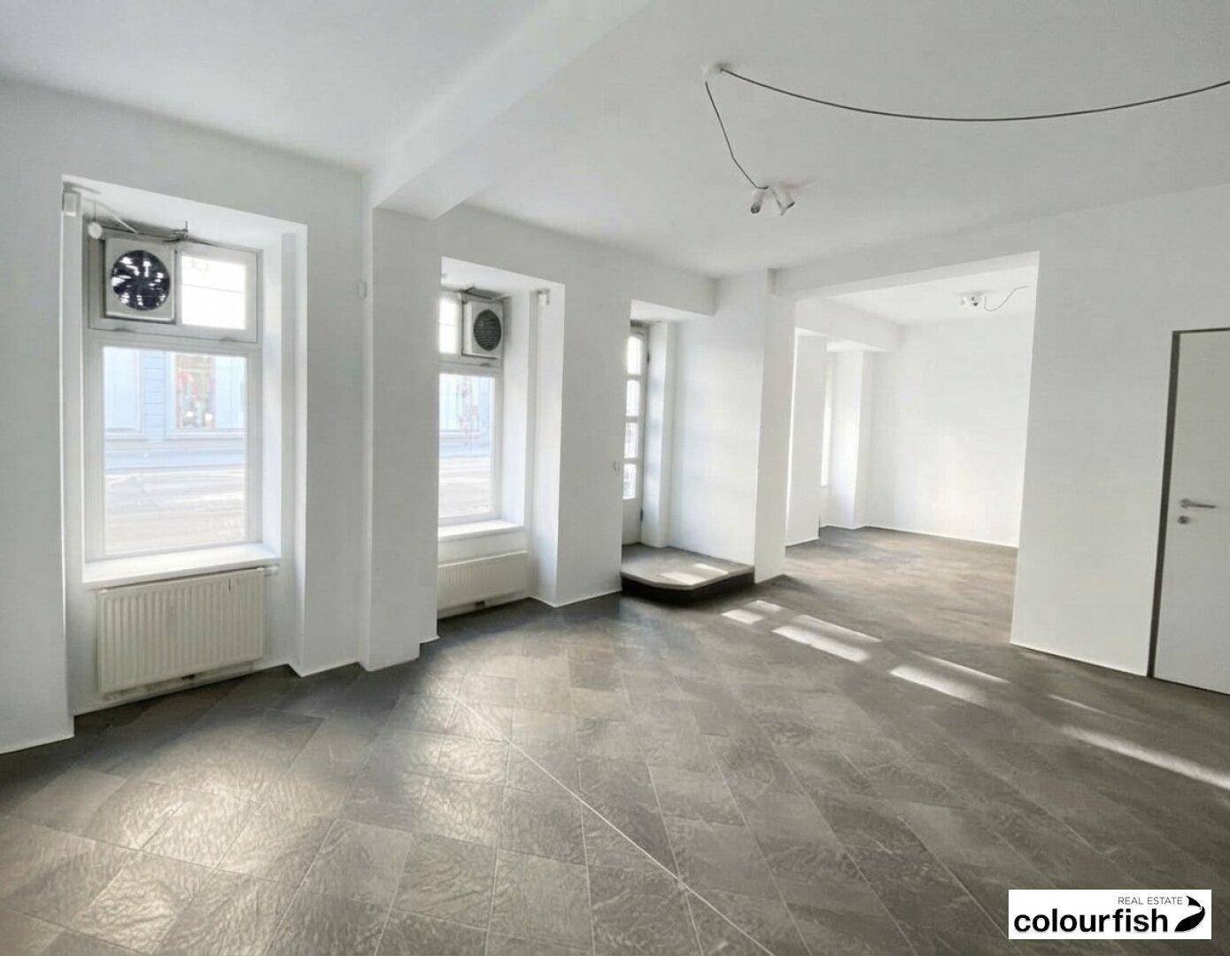 Geräumiger Geschäftsraum mit hellen Wänden, dunklem Fliesenboden und Fenstern mit Lüftungseinheiten.