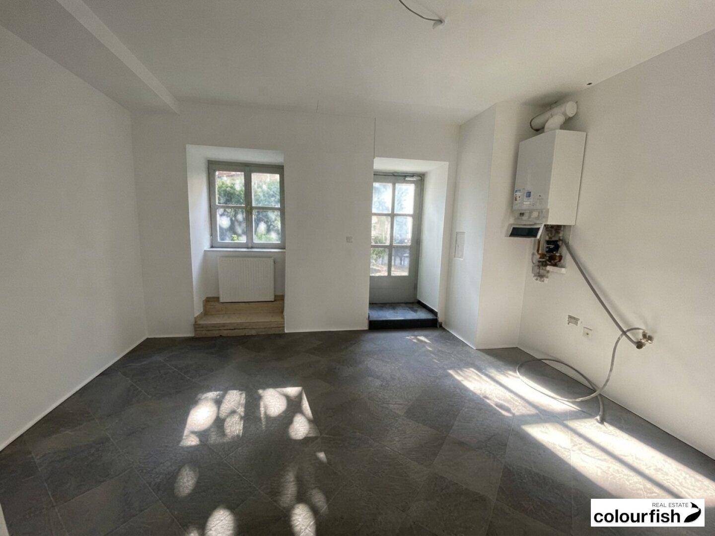 Heller Raum mit weiß gestrichenen Wänden, Fliesenboden und einem sichtbaren Warmwasserbereiter.