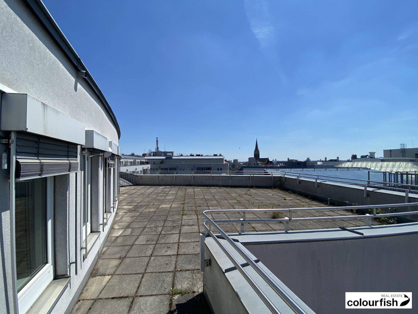 Weitläufige Dachterrasse mit Stadtblick und einem Kirchturm in der Ferne unter strahlend blauem Himmel.