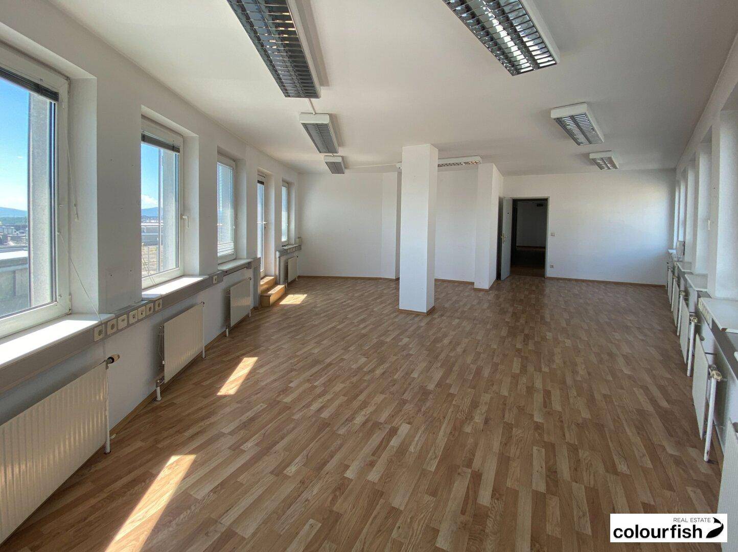 Langer Büroraum mit vielen Fenstern, Säulen und Holzparkettboden, ideal für Großraumbüros.
