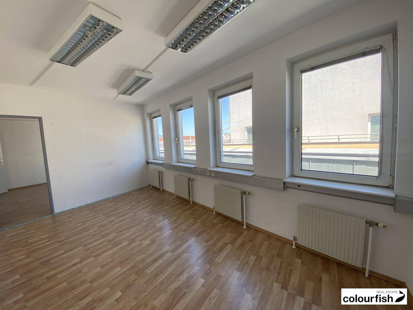 Heller Büroraum mit Holzparkettboden und großen Fenstern, die viel Tageslicht hereinlassen.