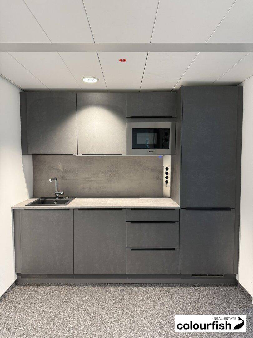 Moderne Küchenzeile mit grauen Schränken, Spüle, Mikrowelle und integriertem Kühlschrank in einem Büro.