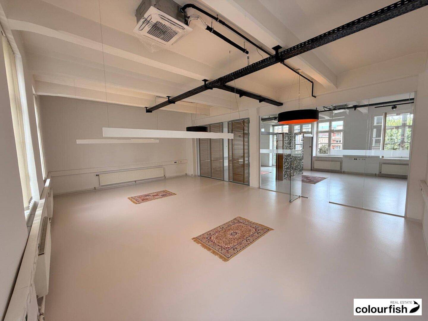 Heller Büroraum mit Teppichen, Glaswänden und sichtbaren Lüftungsanlagen an der Decke.