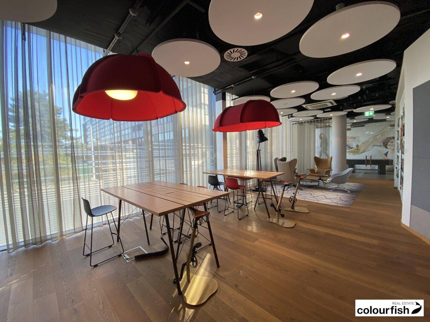 Moderner Bürobereich mit Holzboden, großen Fenstern und auffälligen roten Hängelampen, ideal für kreatives Arbeiten.