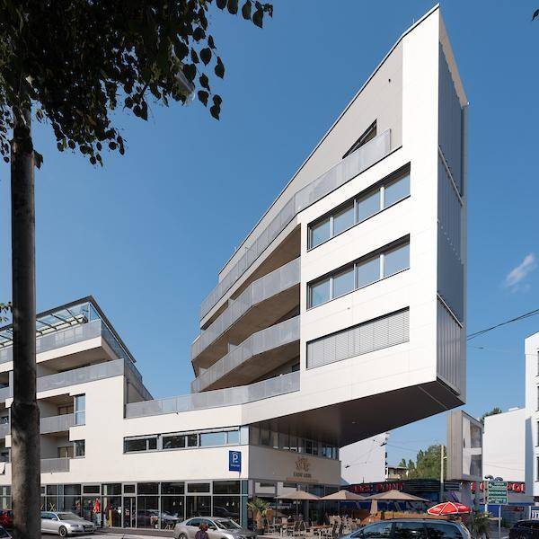 Auffälliges modernes Gebäude mit schräger Fassade, Balkonen und Geschäften im Erdgeschoss.
