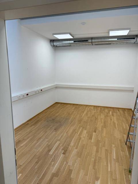 Leerer Büroraum mit hellem Holzparkettboden, weißen Wänden und sichtbaren Lüftungsrohren.