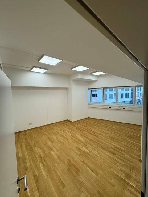 Geräumiger Büroraum mit Holzparkettboden, weißen Wänden und großen Fenstern mit Stadtblick.