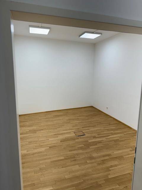 Leerer Büroraum mit hellem Holzparkettboden, weißen Wänden und modernen Deckenleuchten.