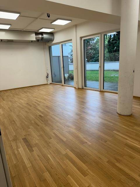 Heller Büroraum mit Holzparkettboden, großen Glastüren und Blick auf eine Grünfläche.