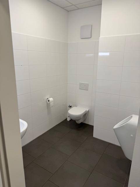 Helle Gästetoilette mit weißen Fliesen, dunklem Boden, WC und Urinal.