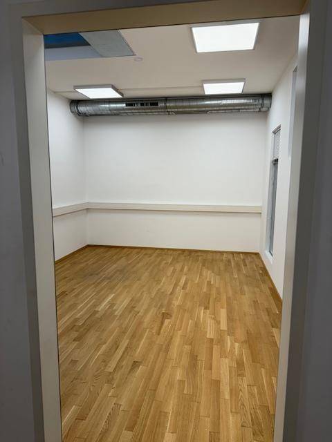 Leerer Büroraum mit hellem Holzparkettboden, weißen Wänden und sichtbaren Lüftungsrohren.