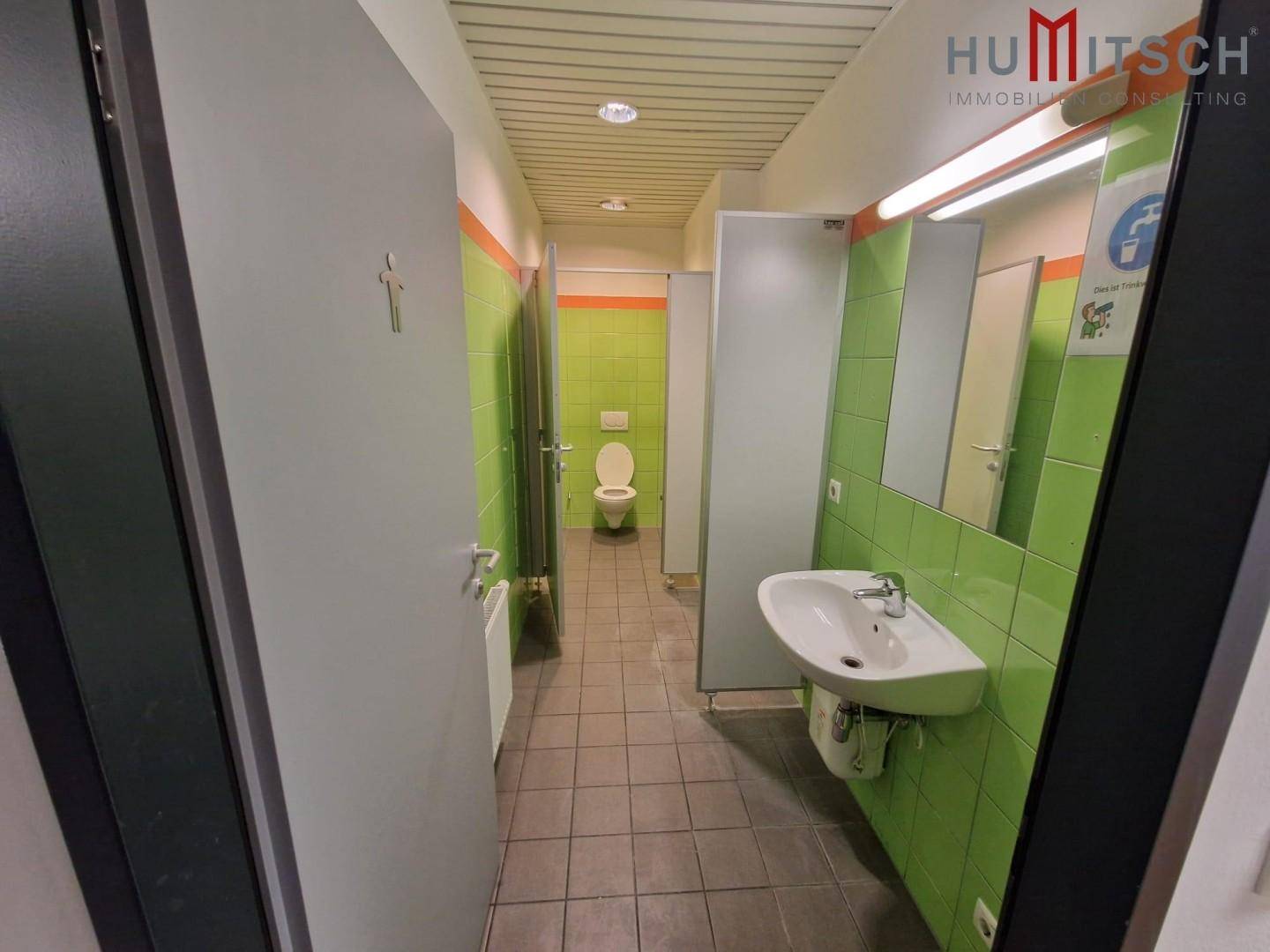 Sanitärbereich mit mehreren Toilettenkabinen, Waschbecken und grünen Fliesen an den Wänden, funktional und sauber.