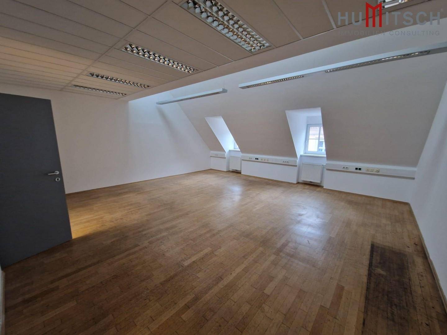 Geräumiges Dachgeschossbüro mit Holzparkettboden, schrägen Wänden und moderner Deckenbeleuchtung.