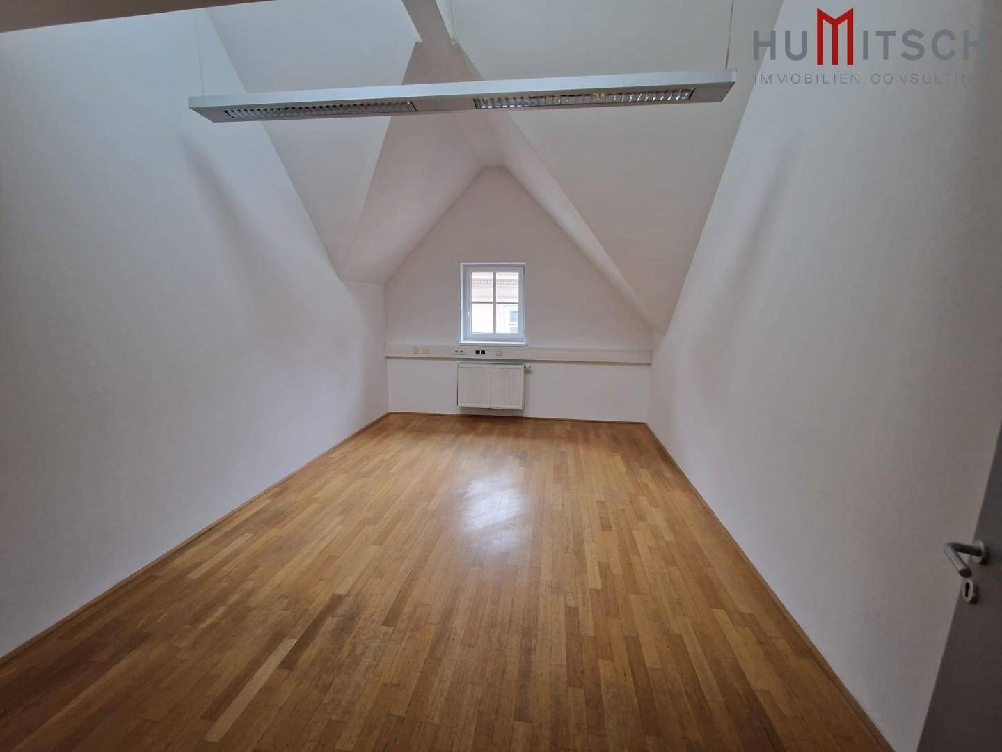 Helles Dachgeschossbüro mit Holzparkettboden und einem einzelnen Fenster in der Giebelwand, gut gepflegt.
