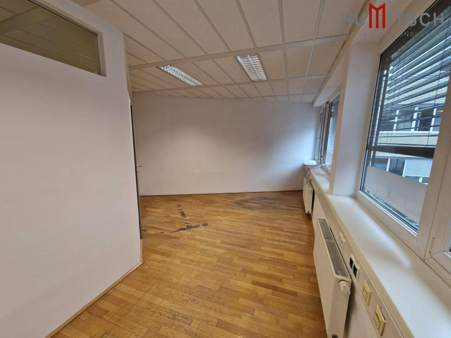 Helles Büro mit Holzparkettboden, Rasterdecke und großen Fenstern, die viel Tageslicht hereinlassen.