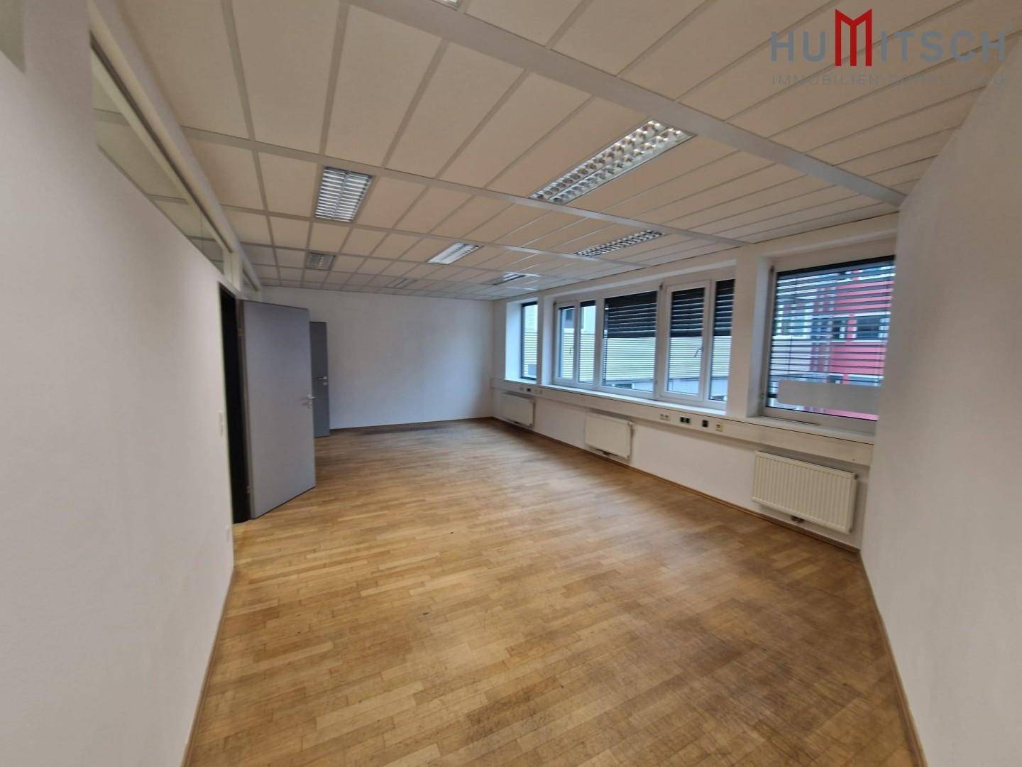 Leeres Büro mit Holzparkettboden, Rasterdecke und großen Fenstern, ideal für individuelle Gestaltung.