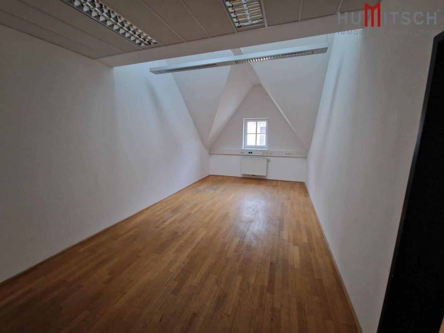 Dachgeschossraum mit Holzparkettboden, schrägen Wänden und einem kleinen Fenster, ideal als ruhiges Büro.