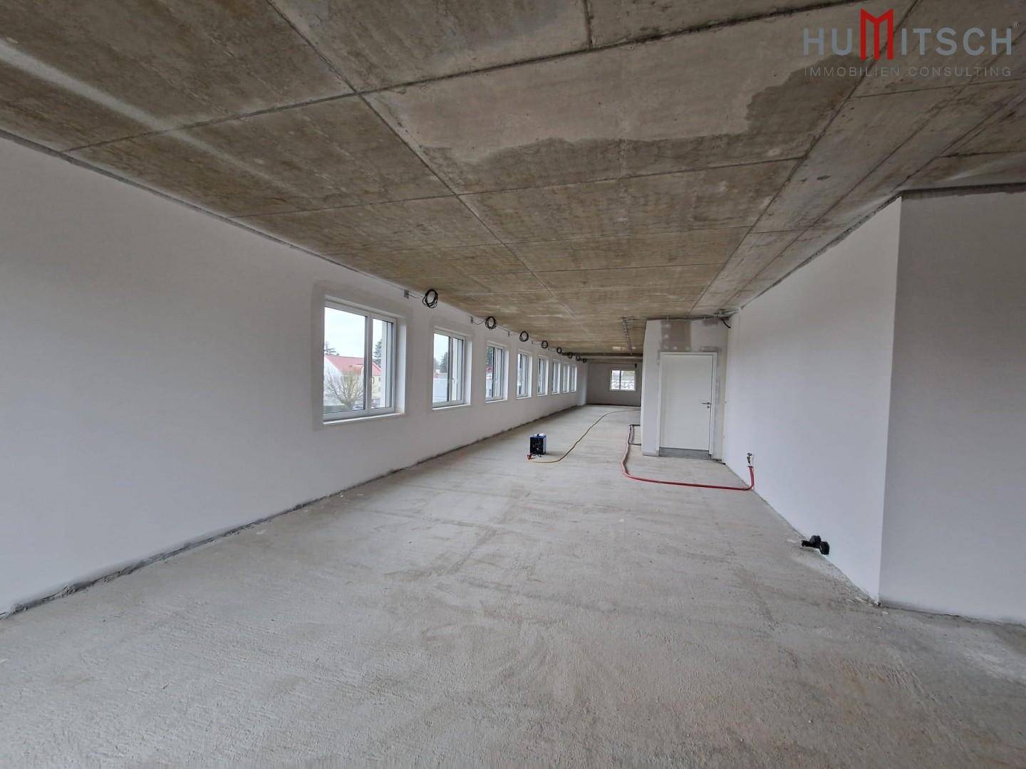 Großer Büroraum im Rohbau mit Betonboden, vielen Fenstern und vorbereiteten Installationen.
