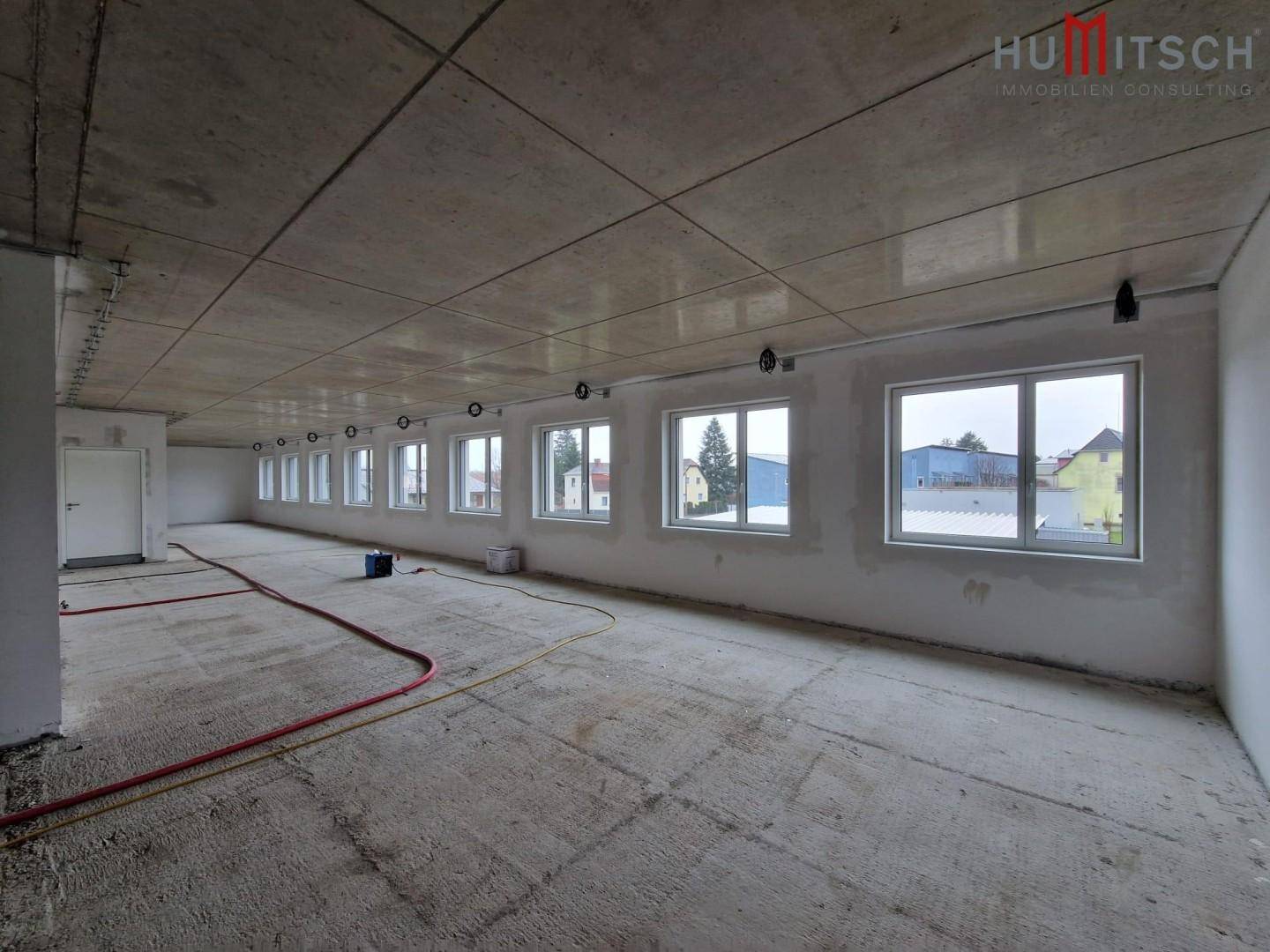 Langer Büroraum im Rohbauzustand mit vielen Fenstern und Betonboden.