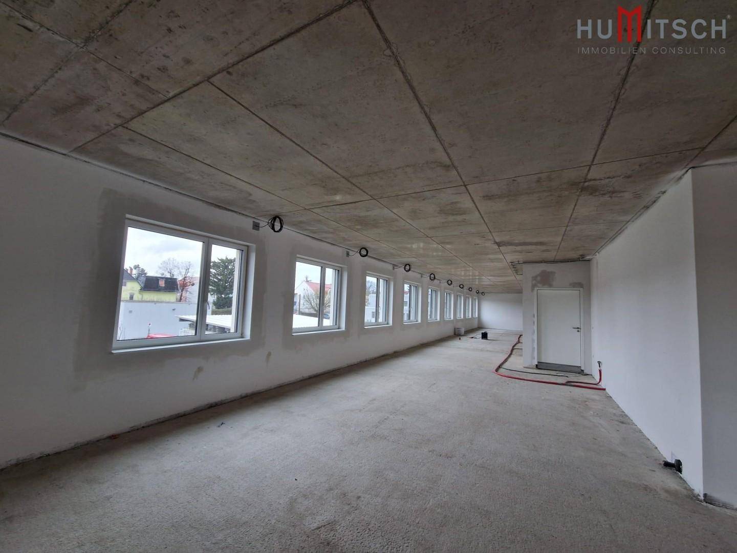 Langer Büroraum im Rohbauzustand mit Betonboden, vielen Fenstern und vorbereiteten Installationen.
