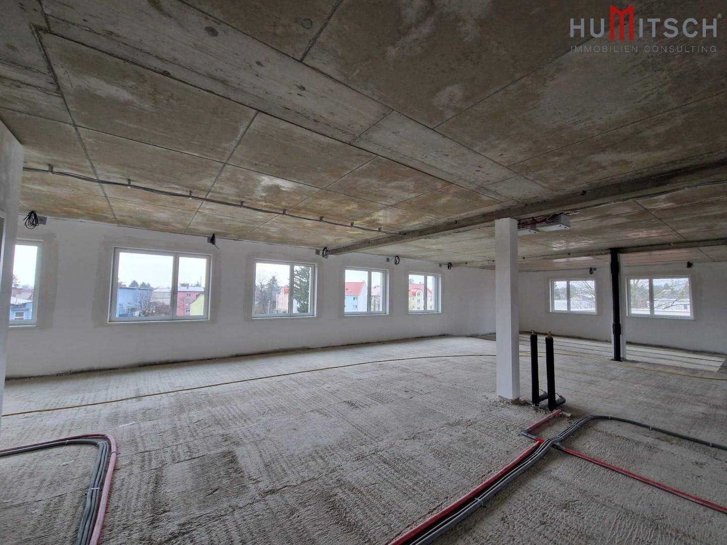 Rohbau eines Büroraums mit Betonboden, sichtbaren Leitungen und vielen Fenstern.